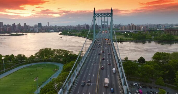 The Triborough bridge with moving cars against river and city panorama. Manhattan skyscrapers at the backdrop of orange sky.