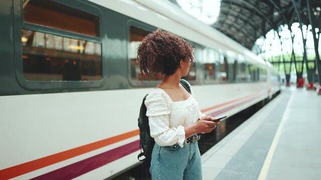 Young woman in glasses with a smartphone walks along the platform next to the train and looks around. Positive woman using mobile phone outdoors in urban background.