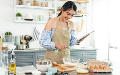 young Asian housewife in kitchen using tablet for searching recipes online cooking the bakery dough homemade.