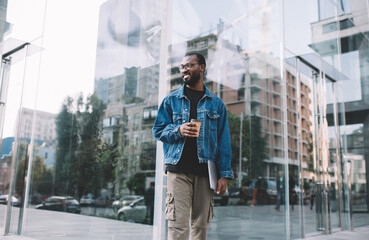 Cheerful dark skinned hipster guy with takeaway caffeine beverage and closed laptop computer smiling outdoors,happy African American digital nomad in spectacles holding coffee to go and modern netbook