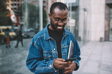 Cheerful male freelancer with closed netbook using cellular gadget for chatting and messaging with...