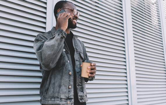 Happy African American hipster guy in optical spectacles for provide eyes correction calling to best friend during coffee break in city, smiling male with takeaway cup using 4g roaming for phoning
