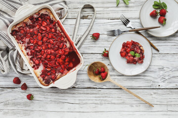 High angle top view of sweet homemade strawberry cobbler or Sonker baked in a red ceramic pan with serving in saucer, apron and wooden spoon over a rustic white wood table. 
