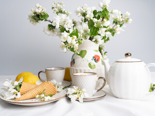 White tea set with tea in cups with yellow lemons and a blossoming apple branch..There are waffle cones on a saucer.