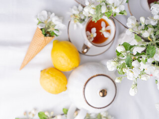 White tea set with tea in cups with yellow lemons and a blossoming apple branch.View from aboveThere are waffle cones on a saucer.
