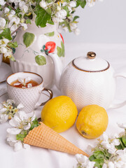 White tea set with tea in cups with yellow lemons and a blossoming apple branch..There are waffle cones on a saucer.