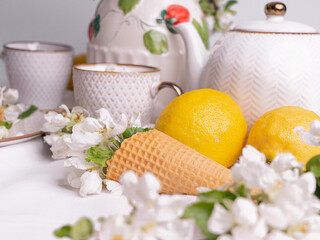 White tea set with tea in cups with yellow lemons and a blossoming apple branch.Close-upThere are waffle cones on a saucer.