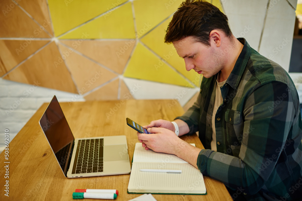 Poster young thoughtful man texting message on mobile phone while sitting at the desk with laptop computer 
