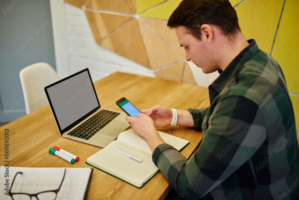 Poster young thoughtful man texting message on mobile phone while sitting at the desk with laptop computer 