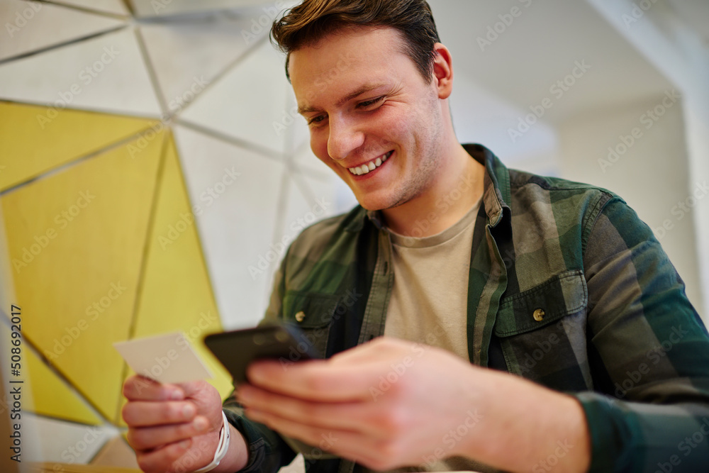 Poster young smiling man texting message on mobile phone while sitting at the desk. using technology. diali