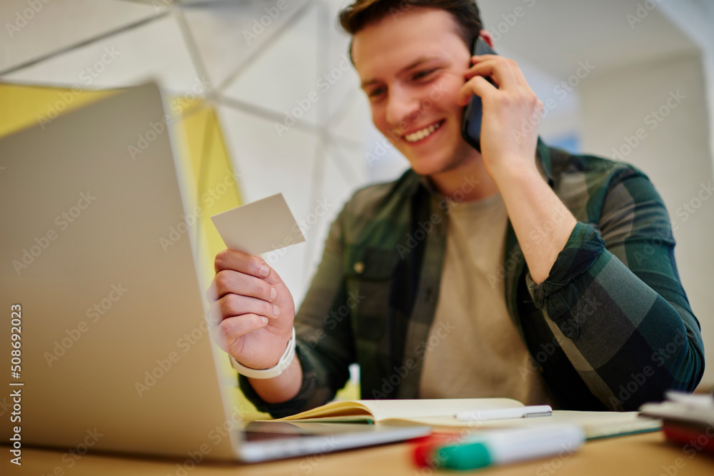 Poster young smiling man calling via mobile phone while sitting at the desk with laptop computer. working o