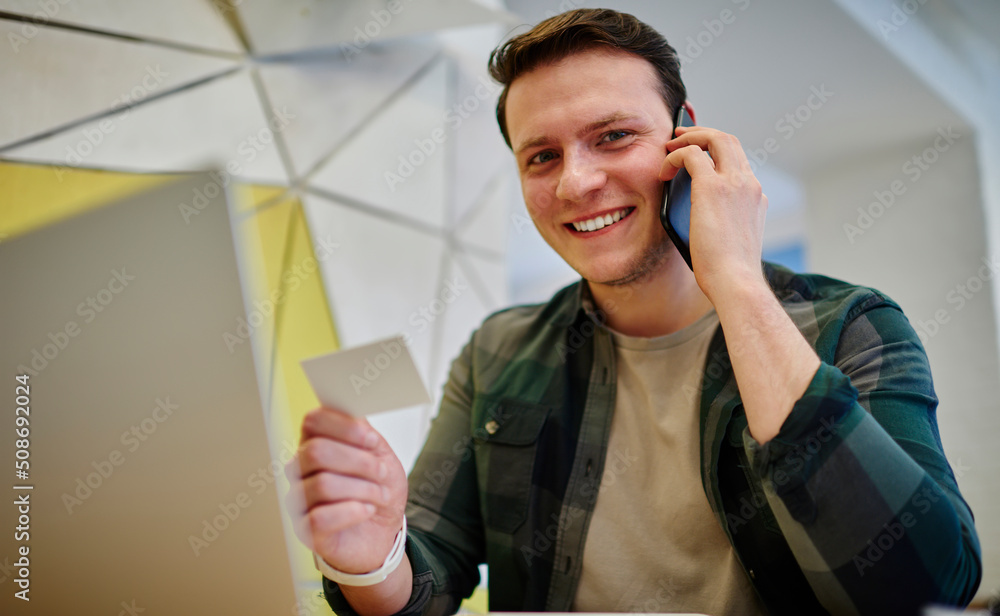 Poster young smiling man calling via mobile phone while sitting at the desk with laptop computer. working o