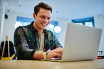 Young smiling man sitting front laptop computer while working online on freelance. Male student learning via netbook. Reading article on website