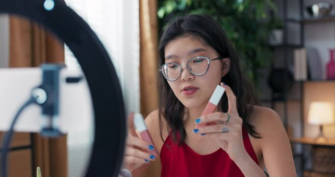 Young vlogger girl showing new kind of lipstick while recording vlog sitting at home. Woman record video indoors, advertising beauty products in front of phone camera.