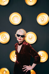 senior stylish elegant woman in sunglasses with glass on lamps dark background. Party, fashion, celebration, anti age concept 