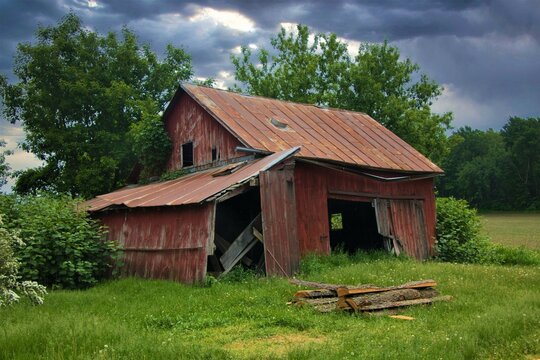 Old, abandoned red farm shed is surrounded by green trees and grasses as a storm approaches in the background.