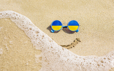 Fototapeta na wymiar Sunglasses with flag of Ukraine on a sandy beach. Nearby is a sea lightning and a painted smile. The concept of a successful vacation in the resorts of Ukraine.