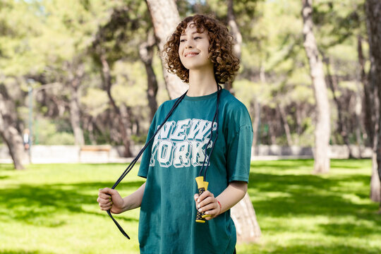 Portrait of young redhead woman wearing green t-shirt standing on city park, outdoor holding a skipping rope on her neck looking away during work out.