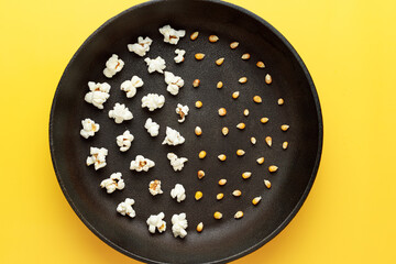 Popcorn in a black pan without a handle, half a pan of corn grains, half of the popcorn. Yellow background. Photography for advertising cinemas, cafes, restaurants, coffee houses. Horizontal photo.