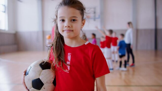 Small l girl standing with ball indoors in gym class, physical education concept