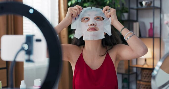 Beauty blogger is live streaming to review makeup products cosmetics on social media while applying fabric beauty mask to face. Influencer uses smartphone on a tripod and a ring light.