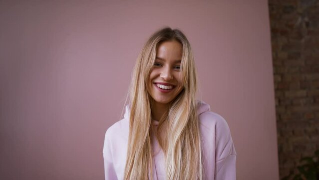 Fashion studio shooting of a happy young blonde woman in hoodie posing over pink background.