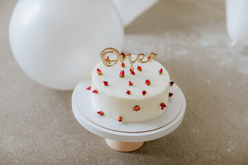White birthday cake for one year celebration is on the floor among white balloons. Focus is on the cake
