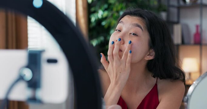 A beautiful Korean blogger demonstrates how to use cosmetics. In front of a mirror and a camera phone, she records a live vlog at home. Facial skin care, applying moisturizer.