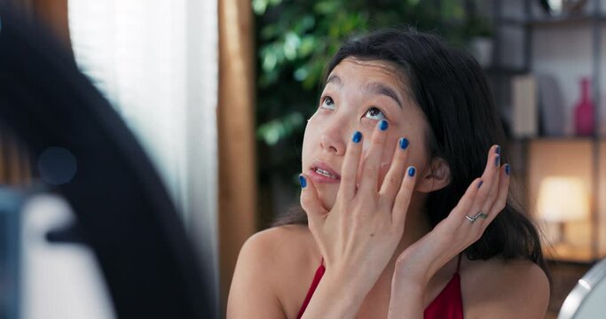 A beautiful Korean blogger demonstrates how to use cosmetics. In front of a mirror and a camera phone, she records a live vlog at home. Facial skin care, applying moisturizer.
