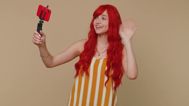 Redhead woman in tank top traveler blogger taking selfie on mobile phone selfie stick, communicating video call online with subscribers. Young ginger girl isolated alone on beige studio background