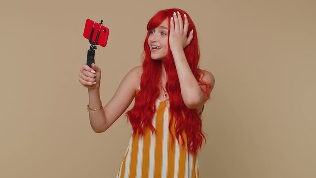 Wow reaction. Impressed redhead woman blogger taking selfie on mobile phone selfie stick, communicating video call online with subscribers. Young girl ginger isolated alone on beige studio background
