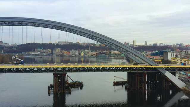 4k aerial footage horizontal span along the unfinished bridge on the river. Construction crane on, boats and barges under, cars driving along road nearby. city view with mixed architecture. Cloudy day