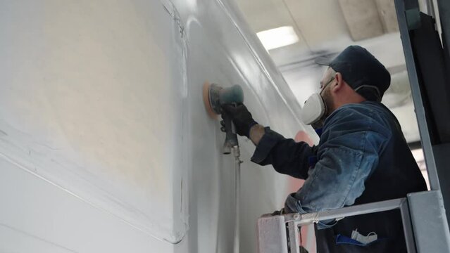 Close-up of a paint line worker in a respirator sanding the car wall before finishing painting using a mechanical sander. Dusty work of a painter in the paint shop of a wagon plant