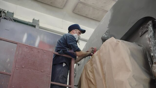 A worker in a cap and a respirator polishes the paintwork of the car using a pneumatic grinder for the subsequent painting of the train. The paint shop of the wagon plant. Depot work.