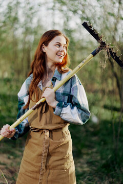 Woman beautifully smiling farmer in work clothes and apron working outdoors in nature and holding a rake to gather grass