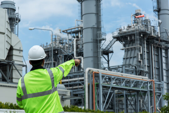 Oil Refinery Factory. Industrial Engineer In Oil Factory. Engineer Wearing PPE Uniform And Helmet Looking Detail Tablet On Hand With Power Plant  On Background.