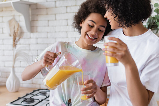 Smiling african american woman pouring orange juice near partner in kitchen