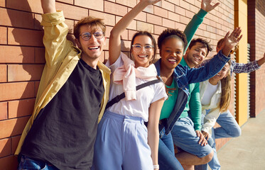 Happy diverse friends having fun together. Group portrait of joyful beautiful young people in casual clothes standing by brick wall on sunny street, smiling, looking at camera and waving hello © Studio Romantic
