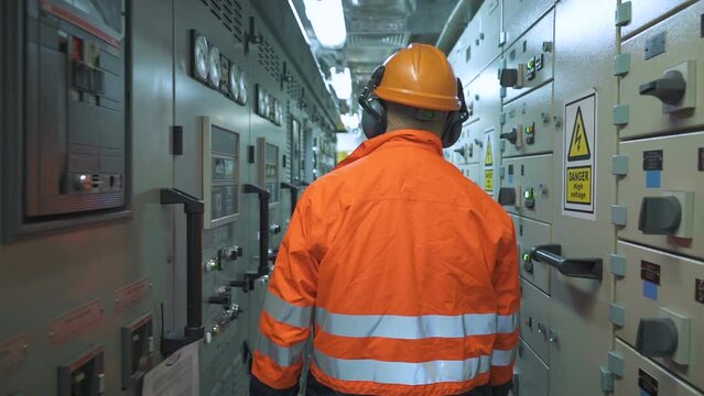 Electrician walks down corridor between electrical panels. Man in orange jacket and helmet with headphones goes. View from back