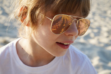 A girl is sitting on a wooden chaise longue on the beach in pink glasses . Reflection in glasses