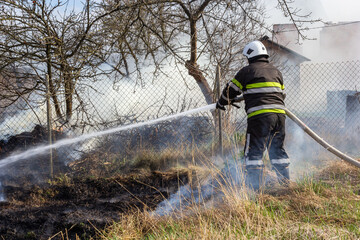 spring fire, burning dry grass near buildings in the countryside. Firefighter extinguishes the...
