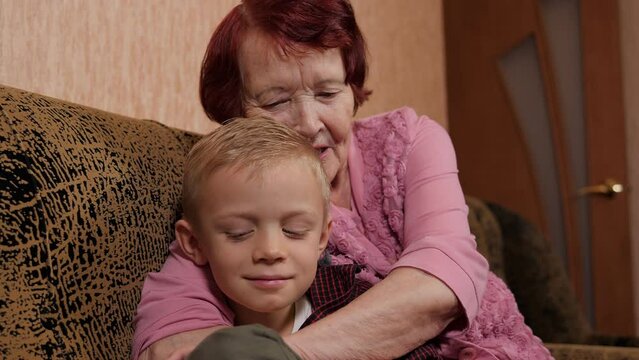Close-up of a happy grandmother kissing her eight-year-old grandson at home sitting on the couch. The grandson came to visit an elderly grandmother.