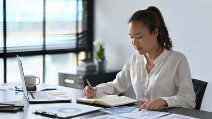 Focused asian businesswoman analyzing financial graph and making notes on notebook