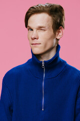 Obraz premium an attractive young man with red hair and freckles on his face looks friendly at the camera winking while standing in a blue zip-up sweater. Vertical portrait on a pink background