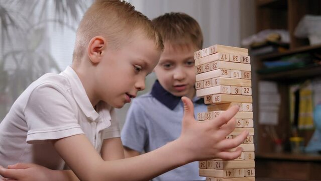 Close-up of two emotional school-age boys playing an exciting wooden blocks board game. The boy takes out a wooden block and the tower starts to wobble.