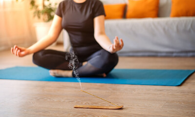 A woman meditates in the lotus position on a turquoise yoga mat on the wooden floor of the house in the living room. Focus on the incense stick and the smoke.