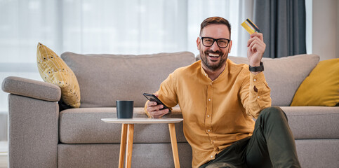 Portrait of cheerful man with credit card using mobile phone in living room at home - obrazy, fototapety, plakaty