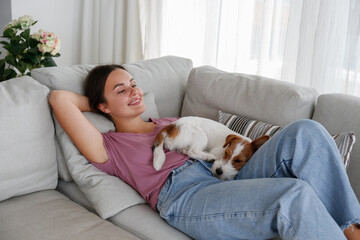 Portrait of young beautiful woman lying on the couch with her adorable wire haired Jack Russel terrier puppy. Loving girl with rough coated pup having fun. Background, close up, copy space.