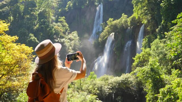 Young woman traveler make photos for social media of amazing exotic waterfalls and tropical nature on her smartphone camera. Happy female tourist with backpack in travel. Wanderlust, tourism concept.