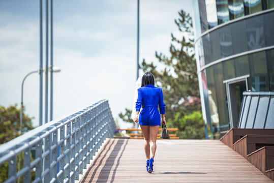 the woman is photographed from behind. The woman walks down the ramp towards the building and is dressed in a blue costume. The woman is in blue shoes and carries a brown handbag. 